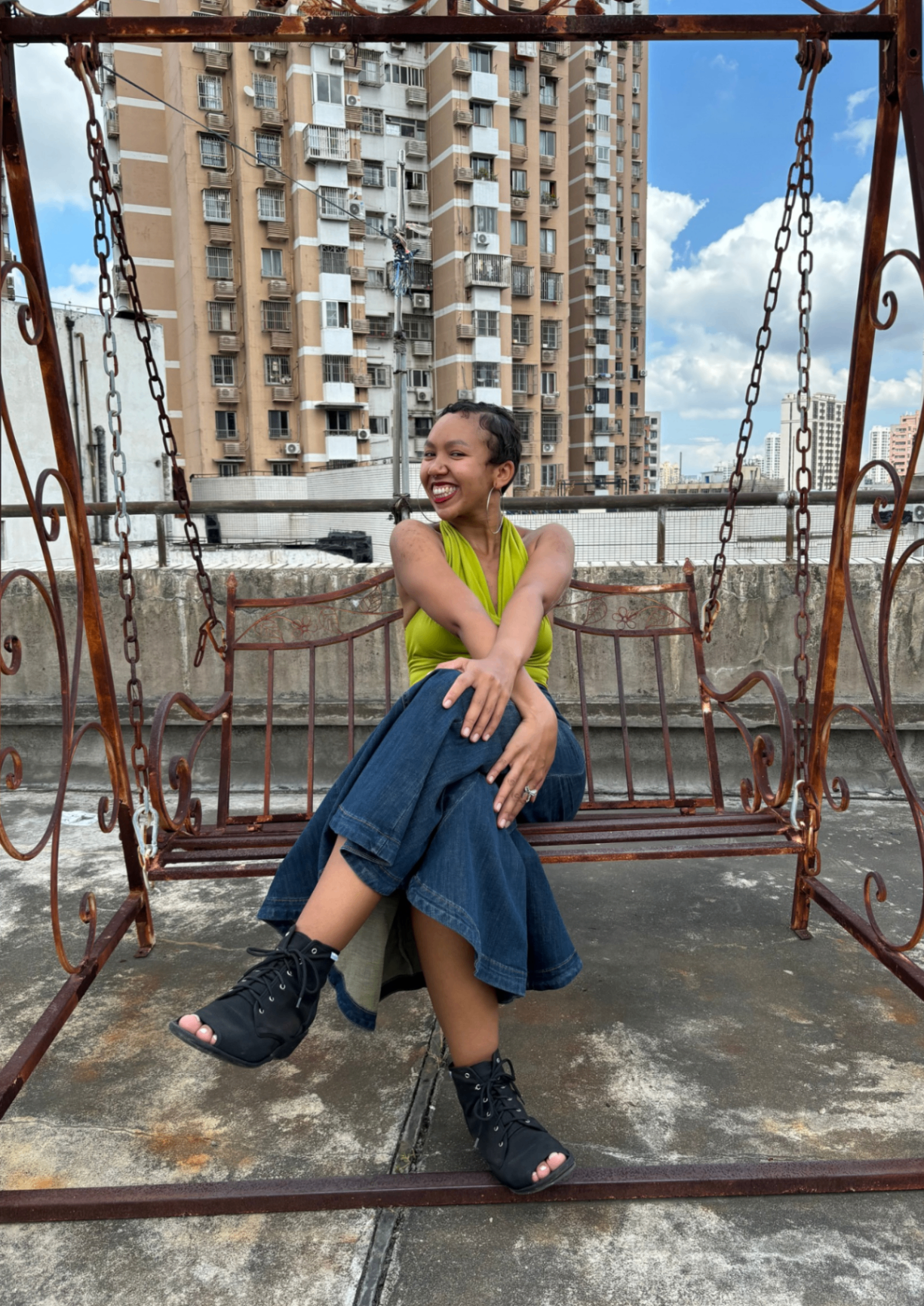 Kanini smiling on a rooftop swing, wearing a green top and denim skirt